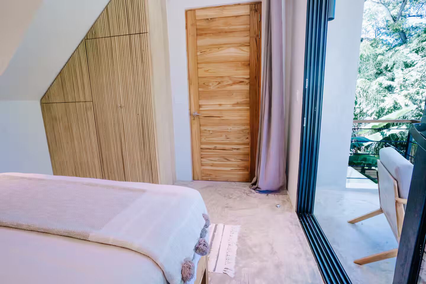 A bedroom with a bed, a wooden door, and a view of a balcony with greenery.