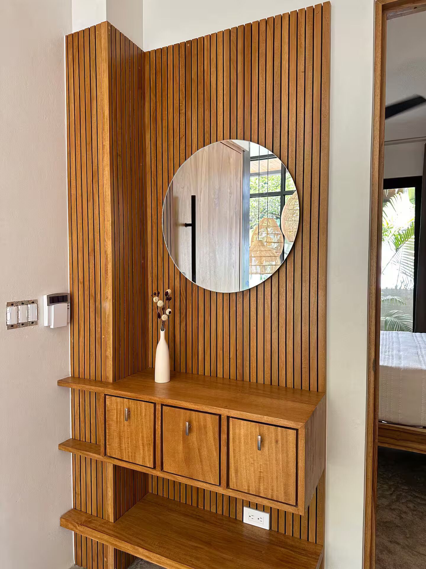 A wooden vanity with a round mirror, a vase with dried flowers, and a built-in shelf.