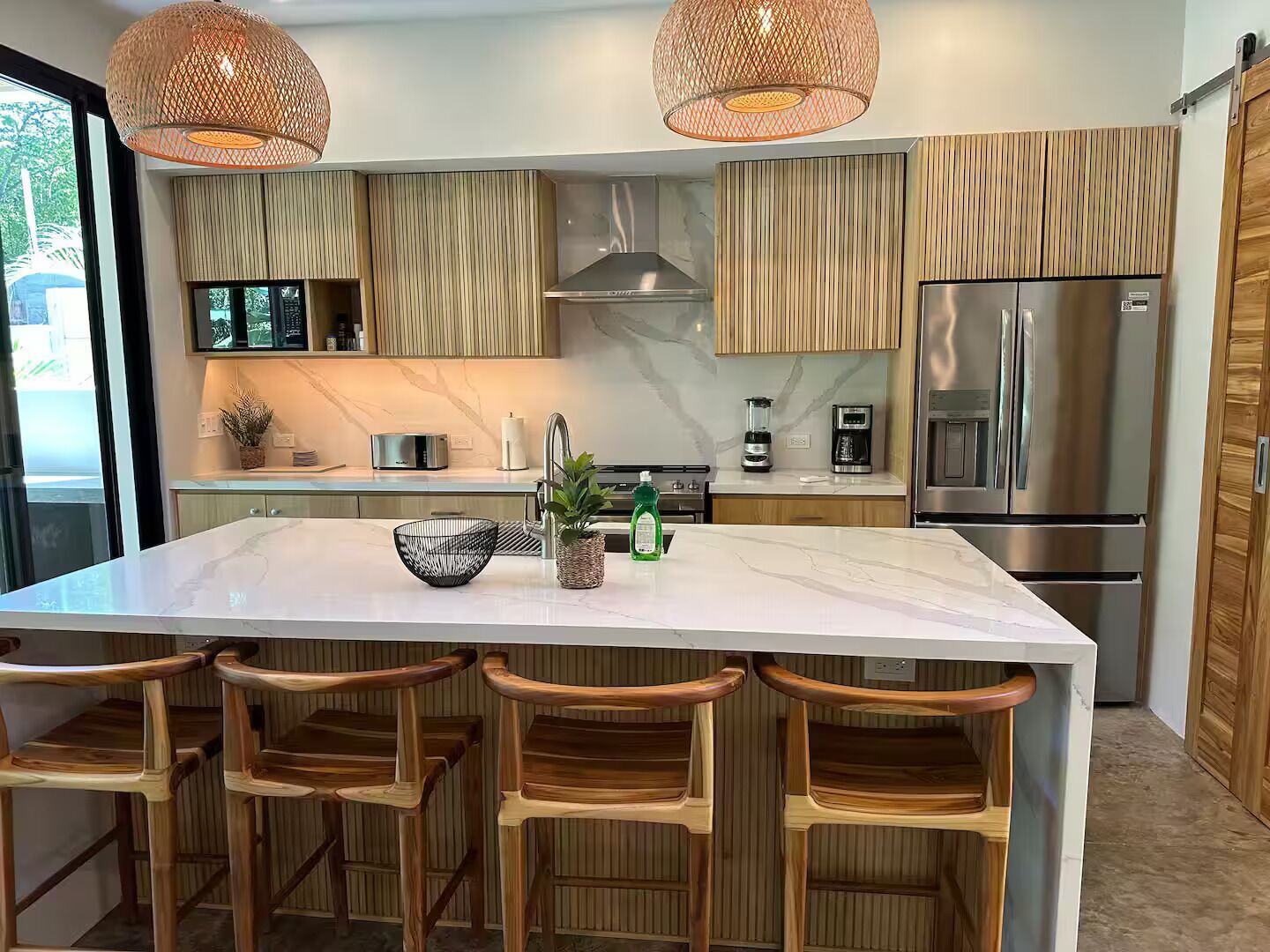 A modern kitchen with a large island, wooden chairs, stainless steel appliances, and pendant lighting.