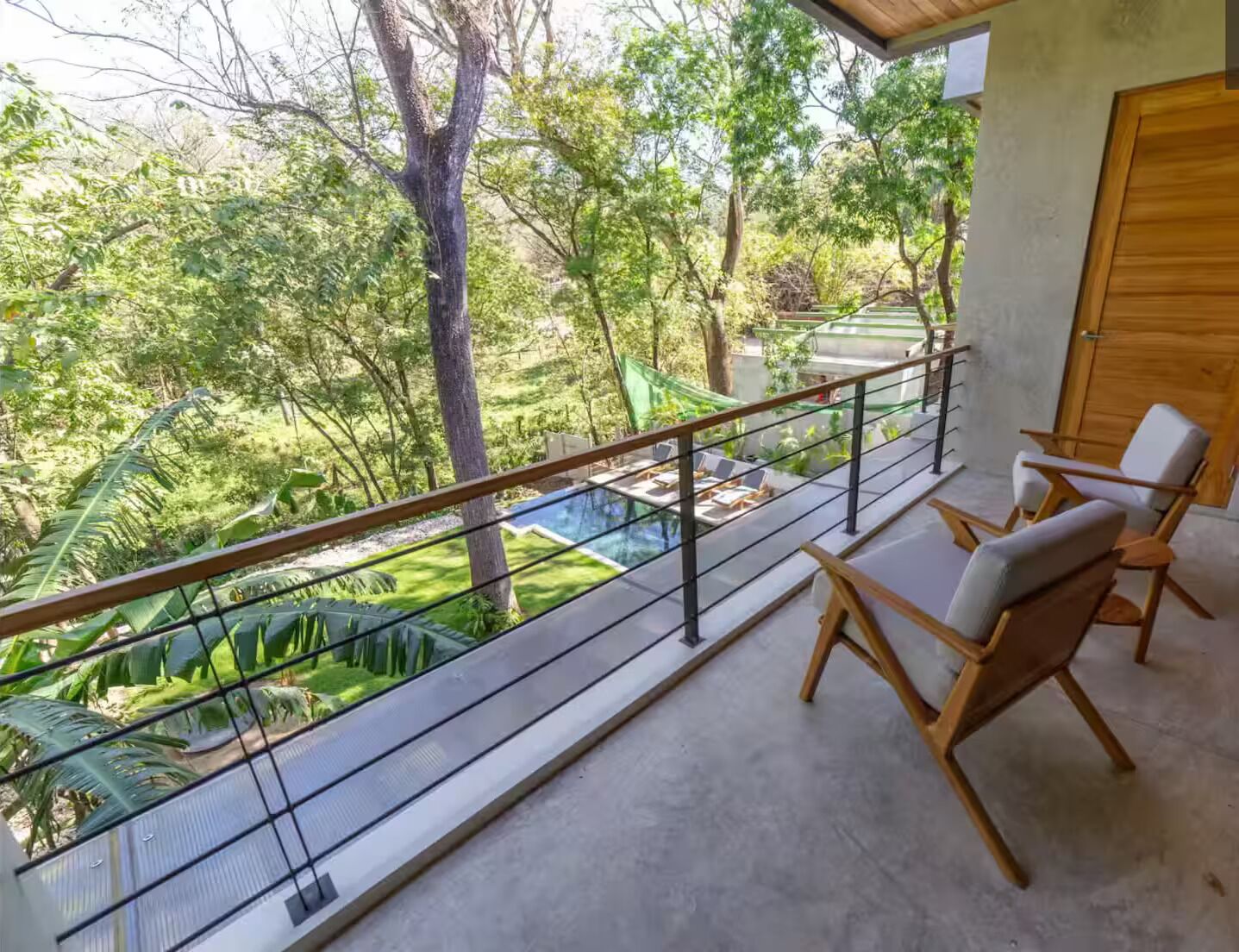 A balcony with a glass railing overlooking a lush garden and a swimming pool.