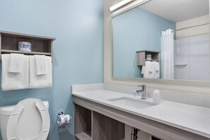 Une salle de bain équipée de toilettes, d’un lavabo et d’un miroir. Des serviettes sont accrochées au mur et une étagère contient des articles de toilette.