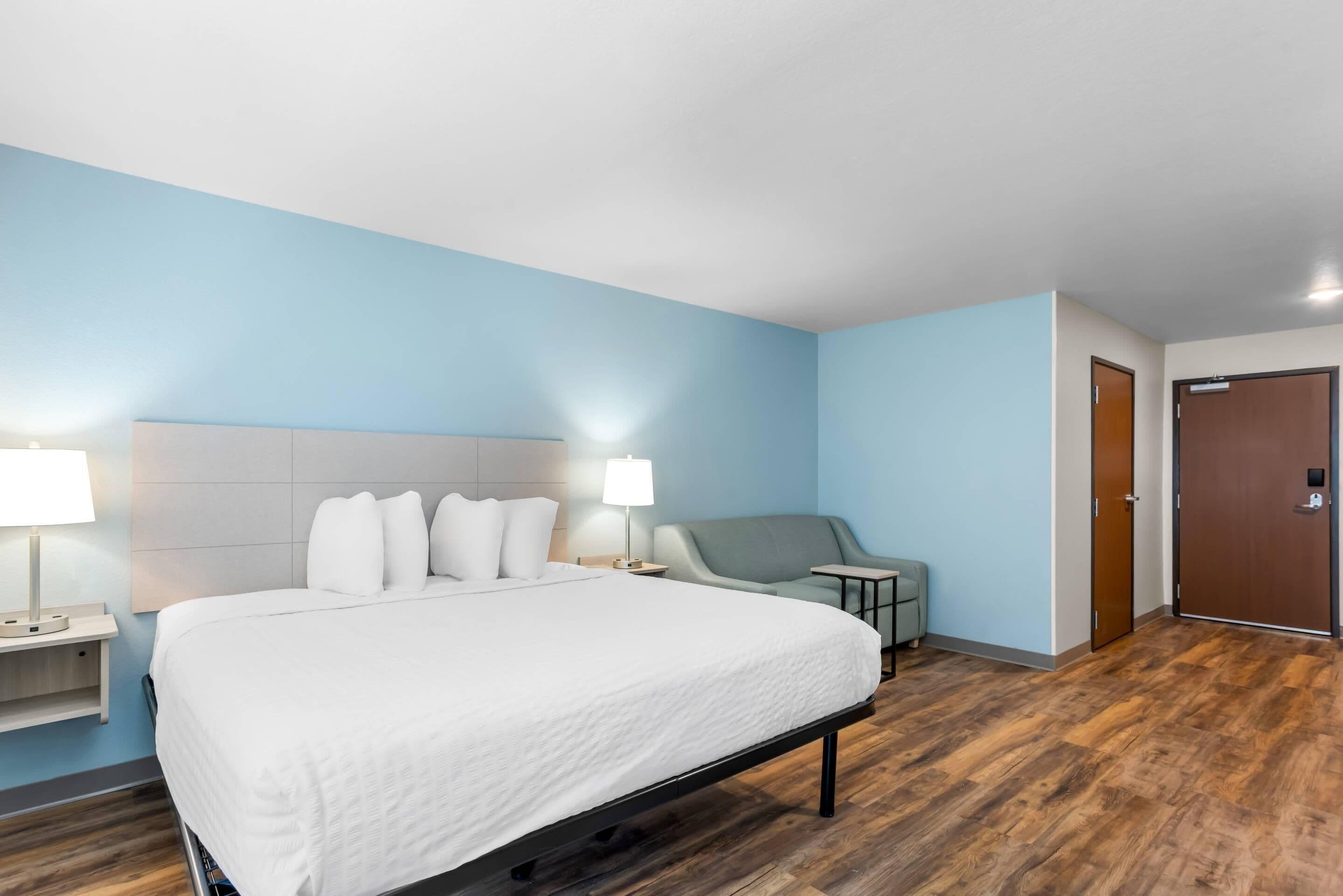 Een hotelkamer met een groot bed, twee bedlampen, een grijze fauteuil, een kleine tafel en een houten vloer.