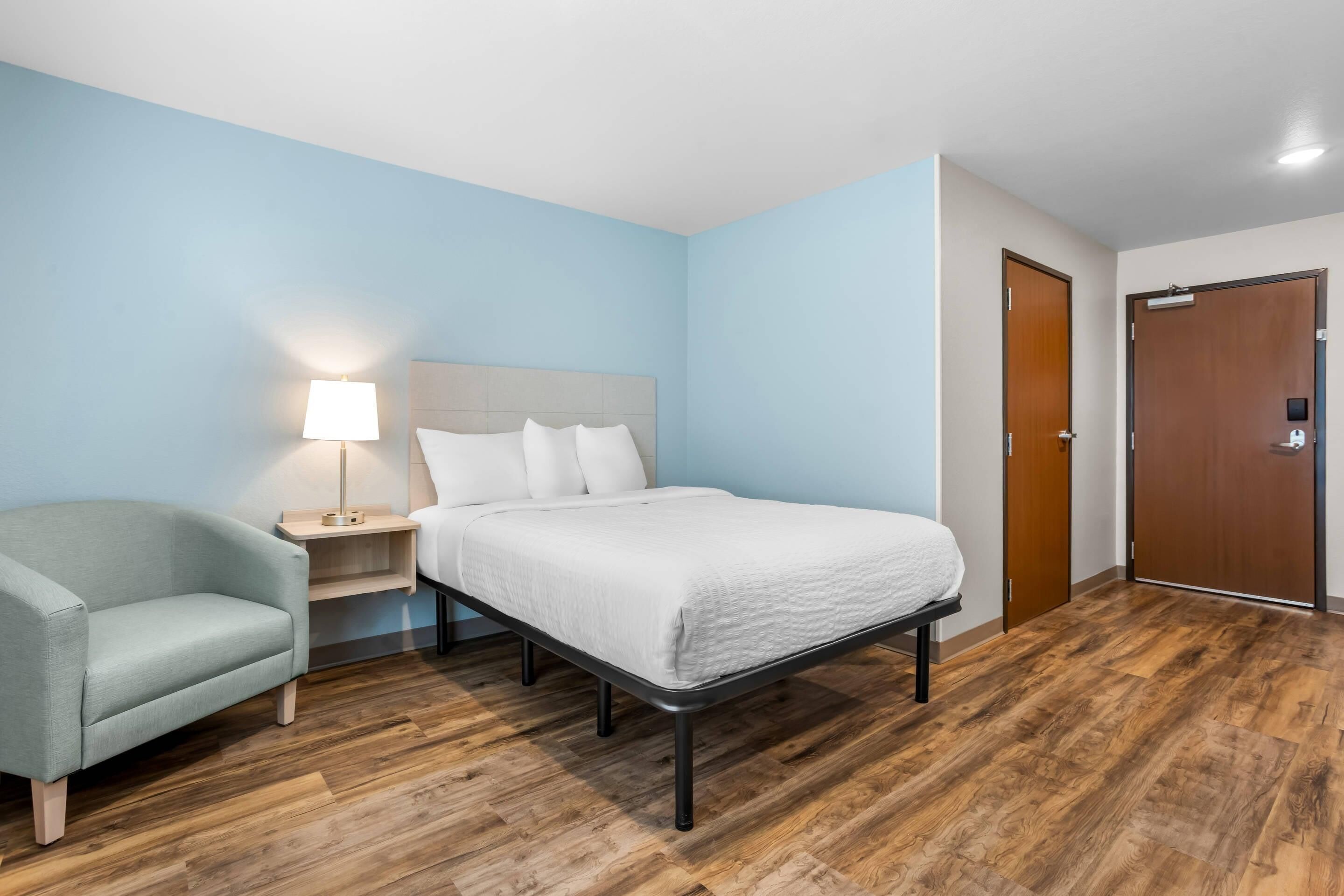 A hotel room with a bed, a nightstand, a lamp, a chair, and wooden flooring.