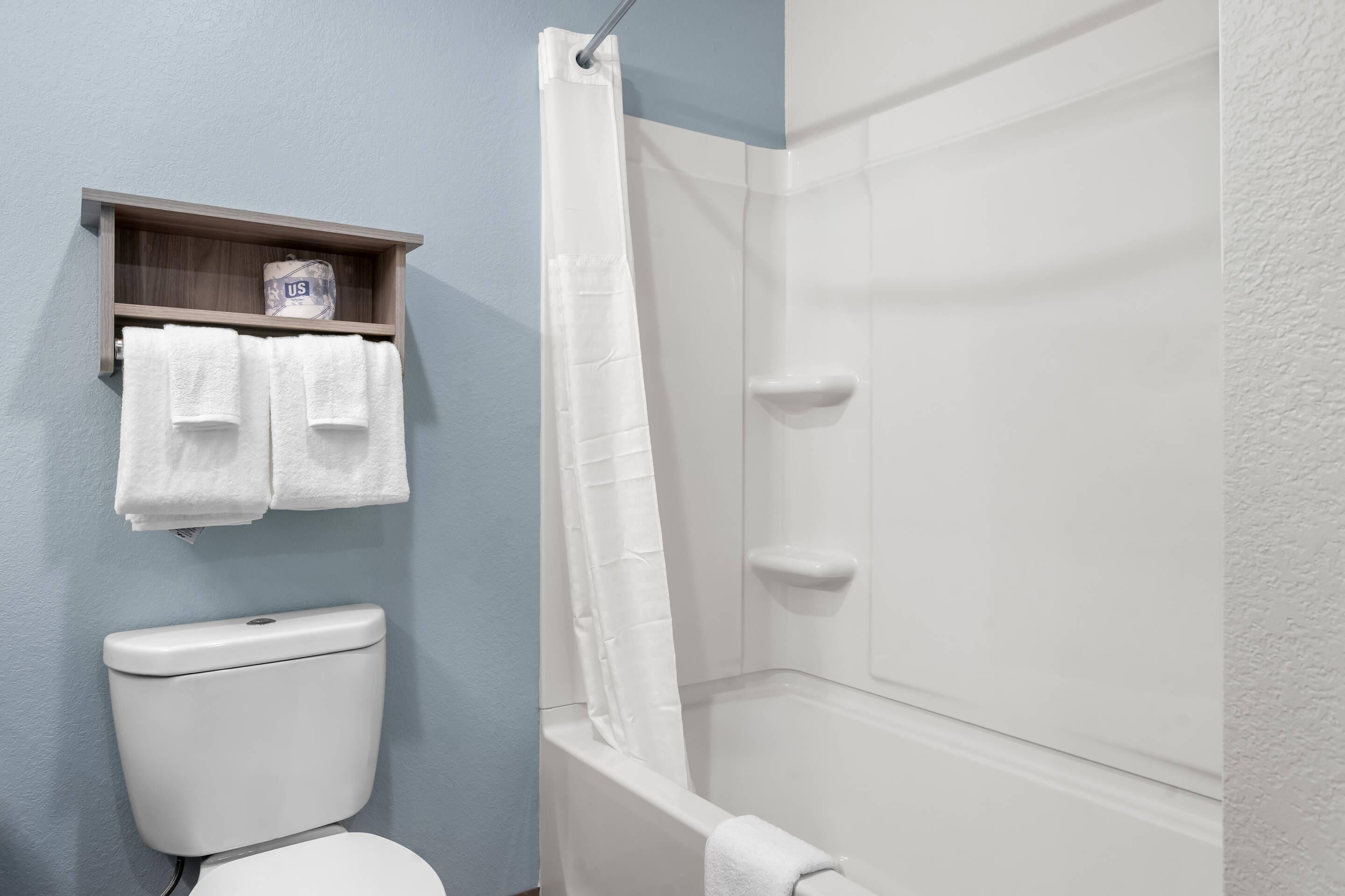 Une salle de bain avec des toilettes, une douche avec un rideau blanc et une étagère sur laquelle se trouve un rouleau de papier toilette.