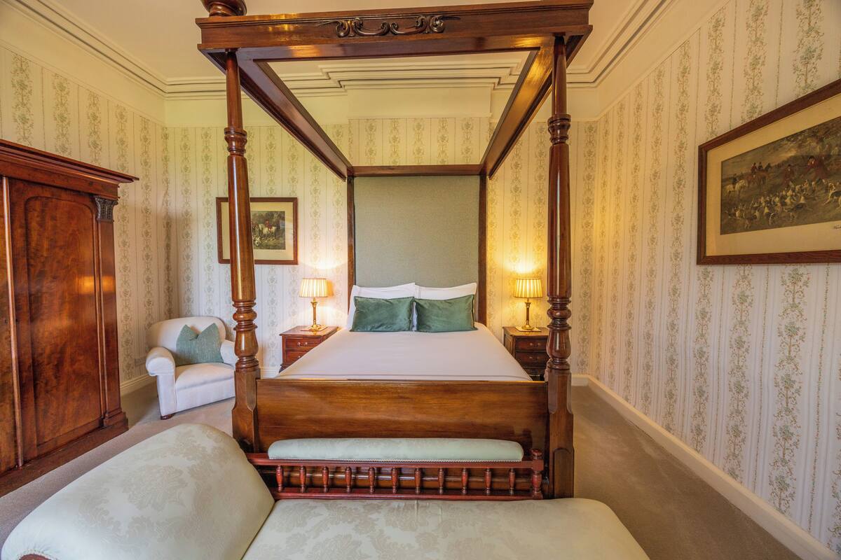 A four-poster bed with a wooden frame, a bench at the foot, a nightstand with a lamp, and a chair with a cushion.