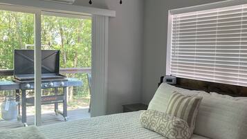 A bedroom with a bed, pillows, a ceiling fan, a window with blinds, and a view of greenery outside.