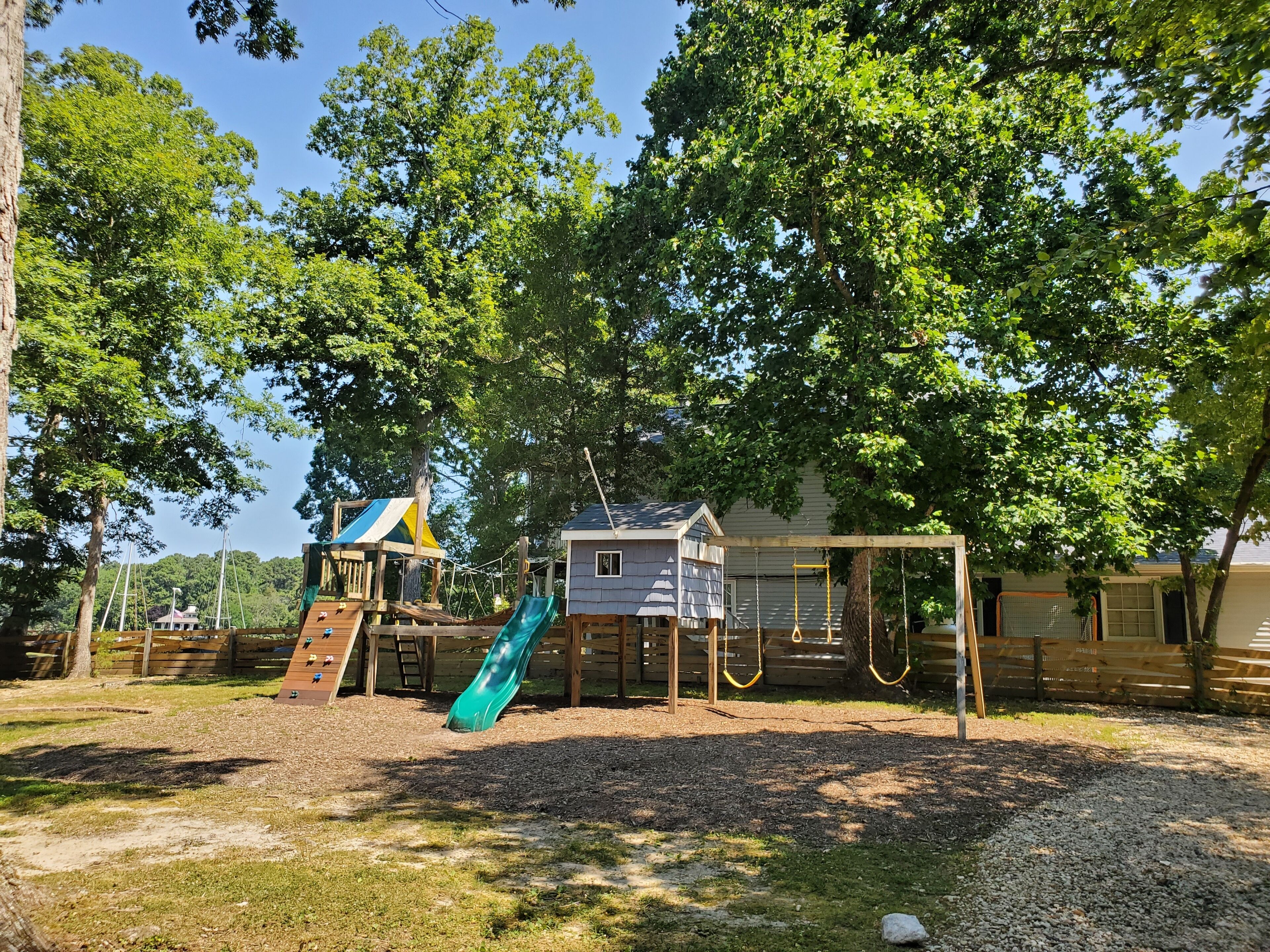 Un terrain de jeux avec un toboggan et des structures d’escalade, entouré d’arbres et avec une maison en arrière-plan.