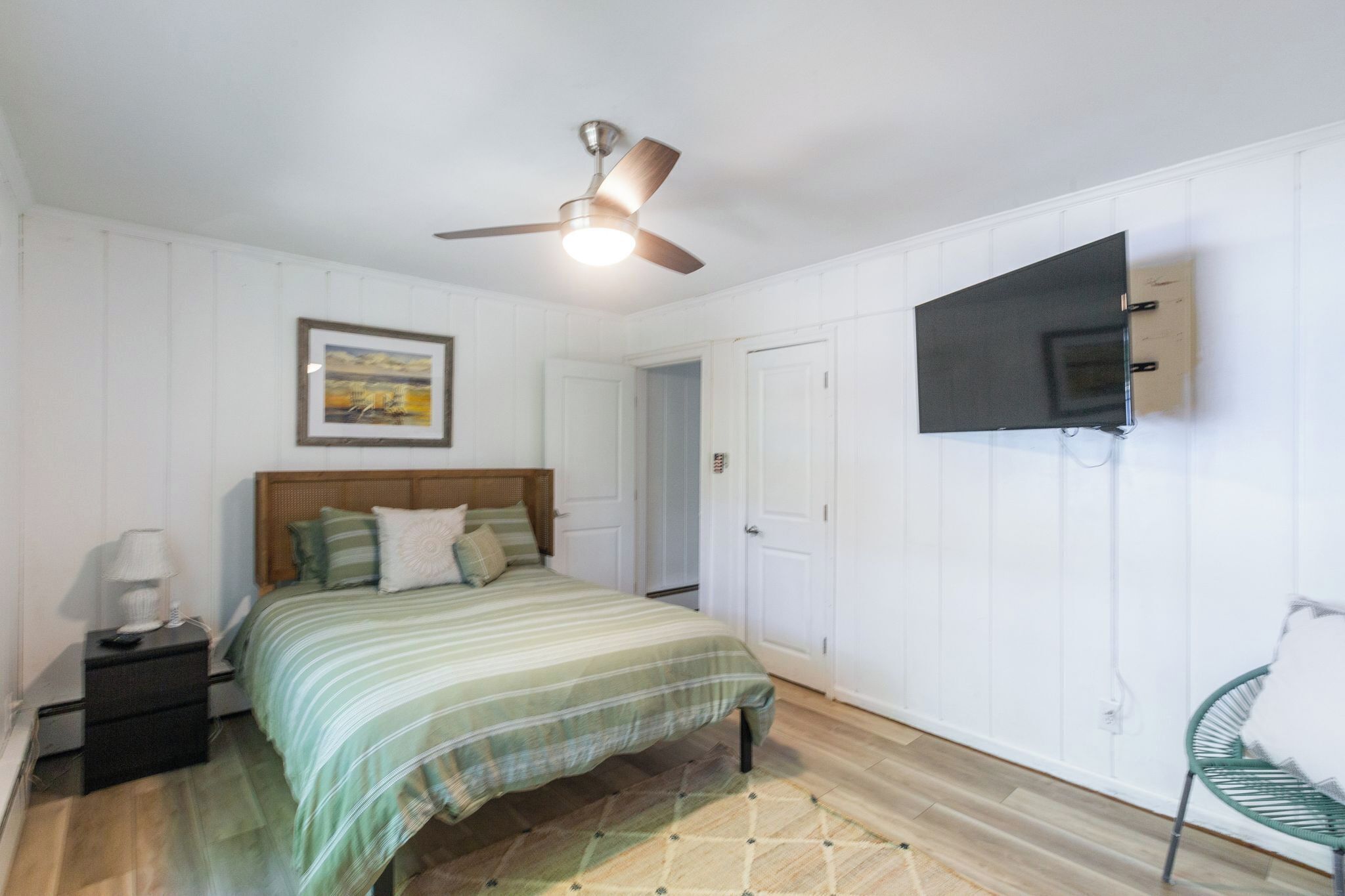 A bedroom with a bed, a nightstand, a ceiling fan, a TV mounted on the wall, and a door.