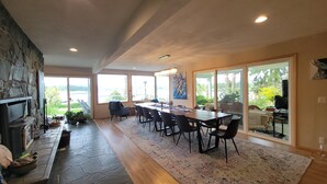 A spacious dining area with a long table and chairs, large windows offering a view of the outdoors, and a stone fireplace.
