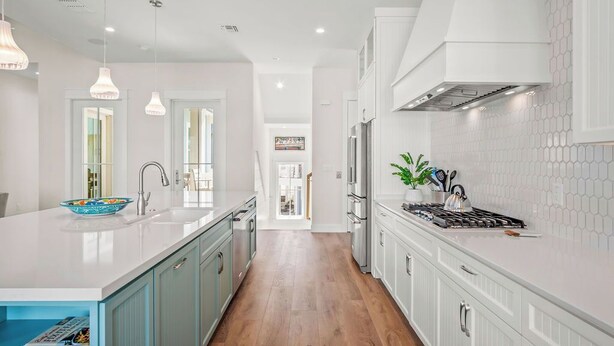 A modern kitchen with white cabinetry, a large island with a sink, pendant lighting, and a view into the dining area. - Fun in the Sun 248 LAK (Port Aransas)