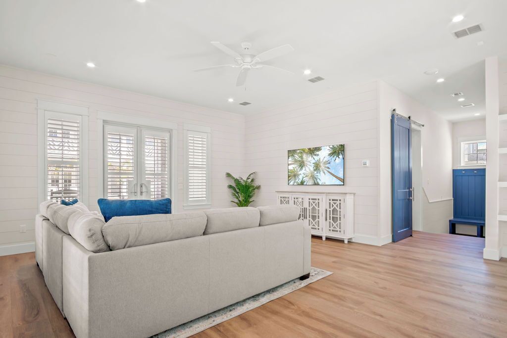 A spacious living room with a beige sofa, white walls, wooden flooring, and a ceiling fan.