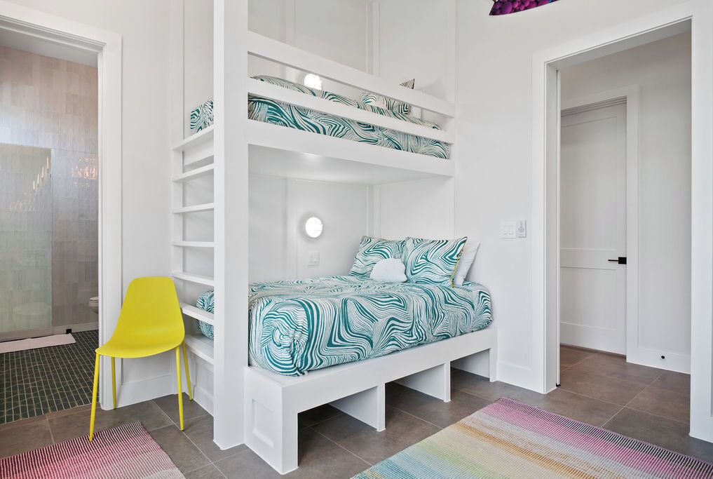 A bunk bed with a patterned bedspread, a yellow chair, and a colorful rug in a room with white walls and a tiled floor.