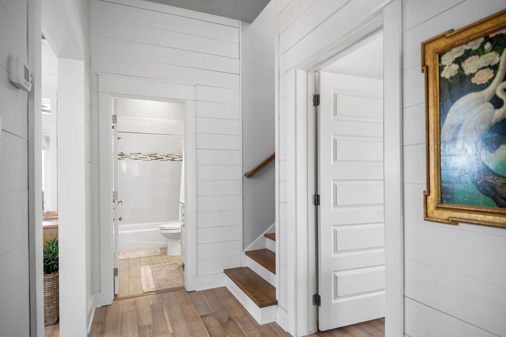 A hallway with white paneled walls, a wooden staircase, and a bathroom with a bathtub and toilet.