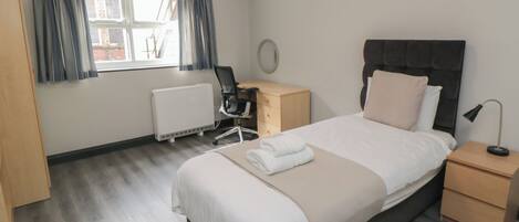 A hotel room with a bed, bedside tables, a desk with a chair, a radiator, and a window with curtains.