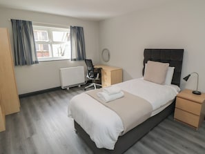 A hotel room with a bed, bedside tables, a desk with a chair, a radiator, and a window with curtains. - The Newborough Apartment 1 (Scarborough)
