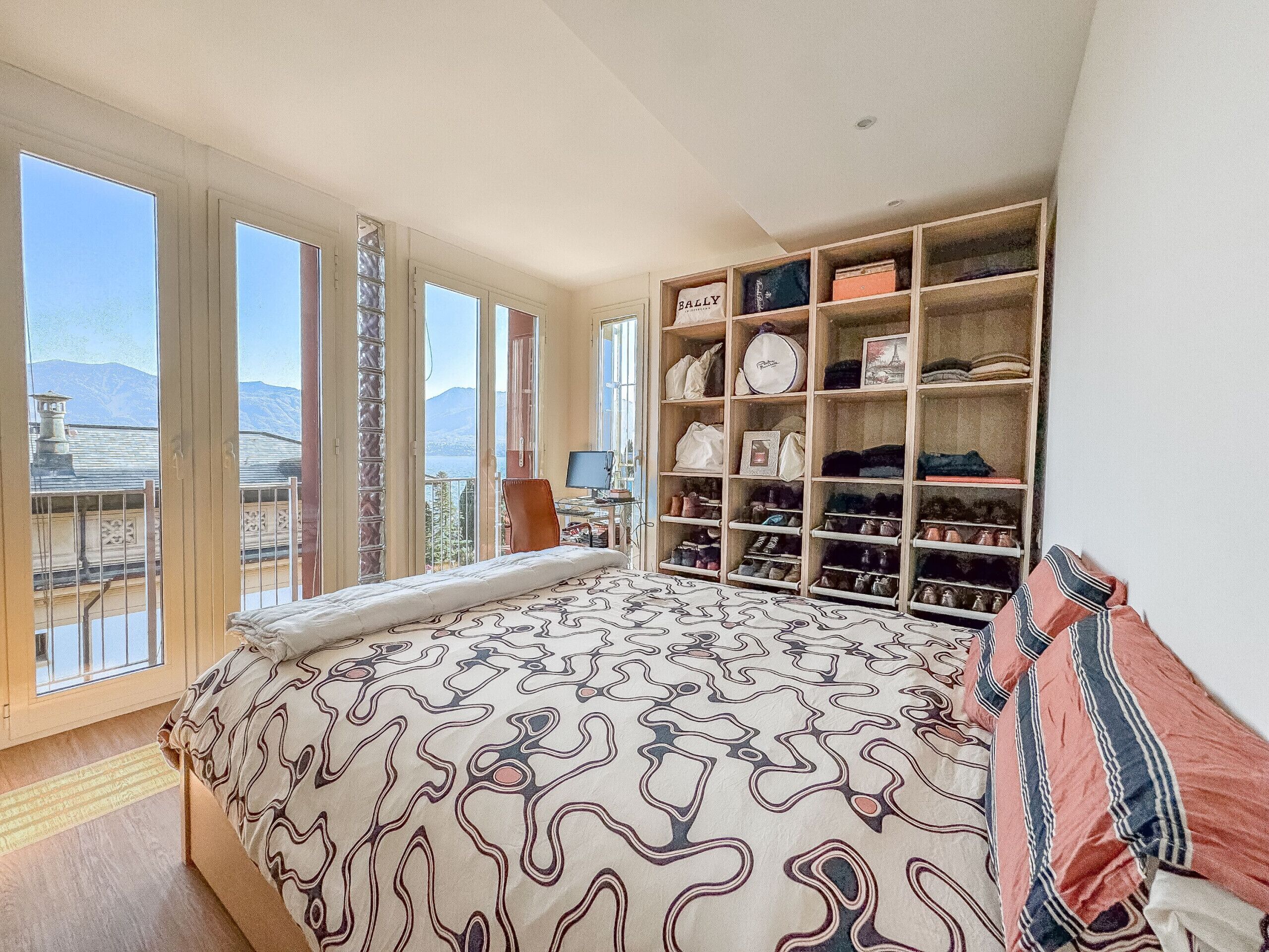 A bedroom with a large bed, a desk with a computer, and a wardrobe with shelves.