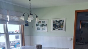 Een keuken met een centraal eiland, witte kasten en een houten vloer. Boven het eiland hangen twee hanglampen en links is een raam met jaloezieën.