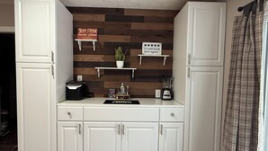 Una cocina con armarios blancos, salpicadero de madera, una máquina de café, una licuadora y un cartel que indica que el bar está abierto.