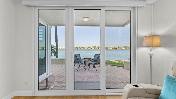 A room with a sliding glass door leading to an outdoor patio with a view of the water and palm trees.