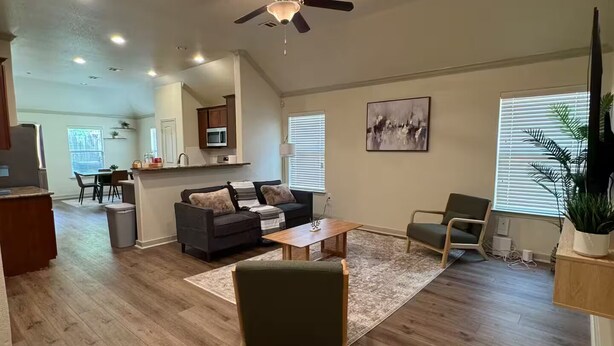 A spacious living area with a ceiling fan, wooden flooring, a sofa, a coffee table, and a dining area in the background.