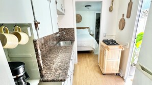 A compact kitchen with a granite countertop, a sink, and a stove. There is a bed visible through an open doorway. - Breezy Bayside Escape — Cottage with Patio in Heart of Mission Beach (San Diego)