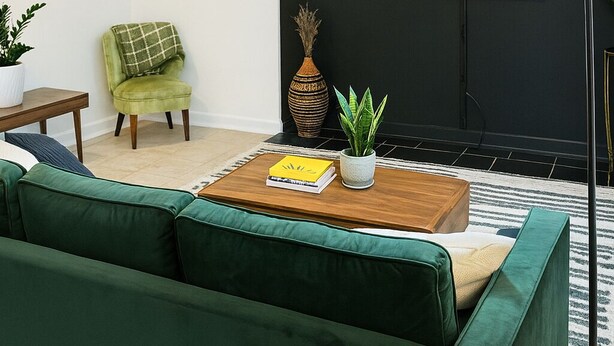 Un salon moderne avec un canapé vert, une petite table basse en bois sur laquelle sont posés un livre et une plante, un fauteuil vert, un grand lampadaire et un téléviseur à écran plat fixé à un mur foncé.