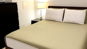 Un lit bien fait, avec une couvre-lit verte et deux oreillers, une table de chevet avec une lampe, et une commode sur la gauche.