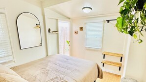 A bedroom with a bed, a wooden floor, a hanging plant, a mirror, and a window with blinds. - Cozy Cottage Between the Beach & Bay East – Walk to Water! (San Diego)