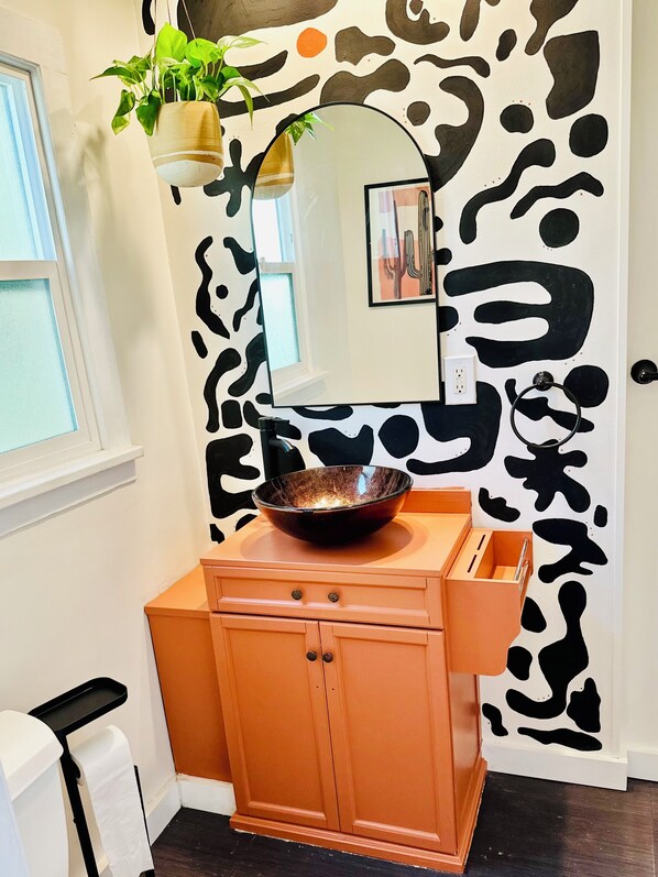 Un baño con una pared blanca que presenta un diseño abstracto negro, un mueble de madera con un cuenco encima, una planta colgante y un espejo sobre el mueble.