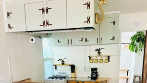 A modern kitchen with a central island, two bar stools, a gas stove, a coffee maker, and decorative wall shelves. - Cozy Cottage Between the Beach & Bay East – Walk to Water! (San Diego)