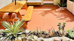 A patio with a wooden table and bench, surrounded by potted plants and a white picket fence. - Cozy Cottage Between the Beach & Bay East – Walk to Water! (San Diego)