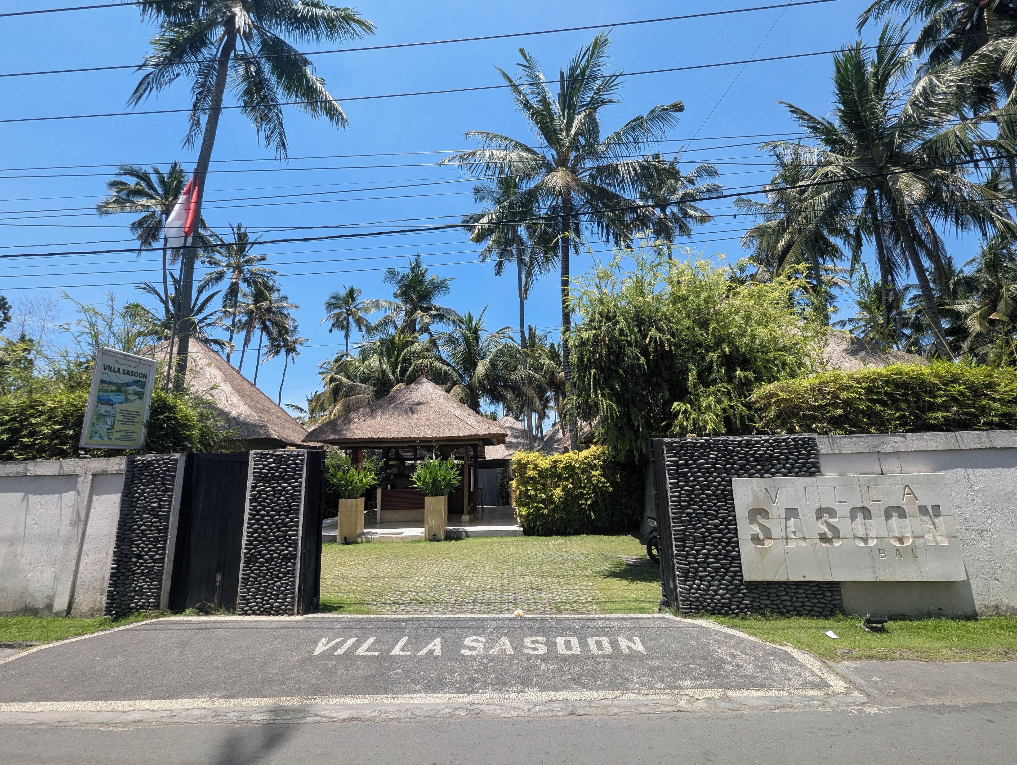 Una entrada con portón y señalización 'Villa Sasoon', una estructura de techo de paja y palmeras.