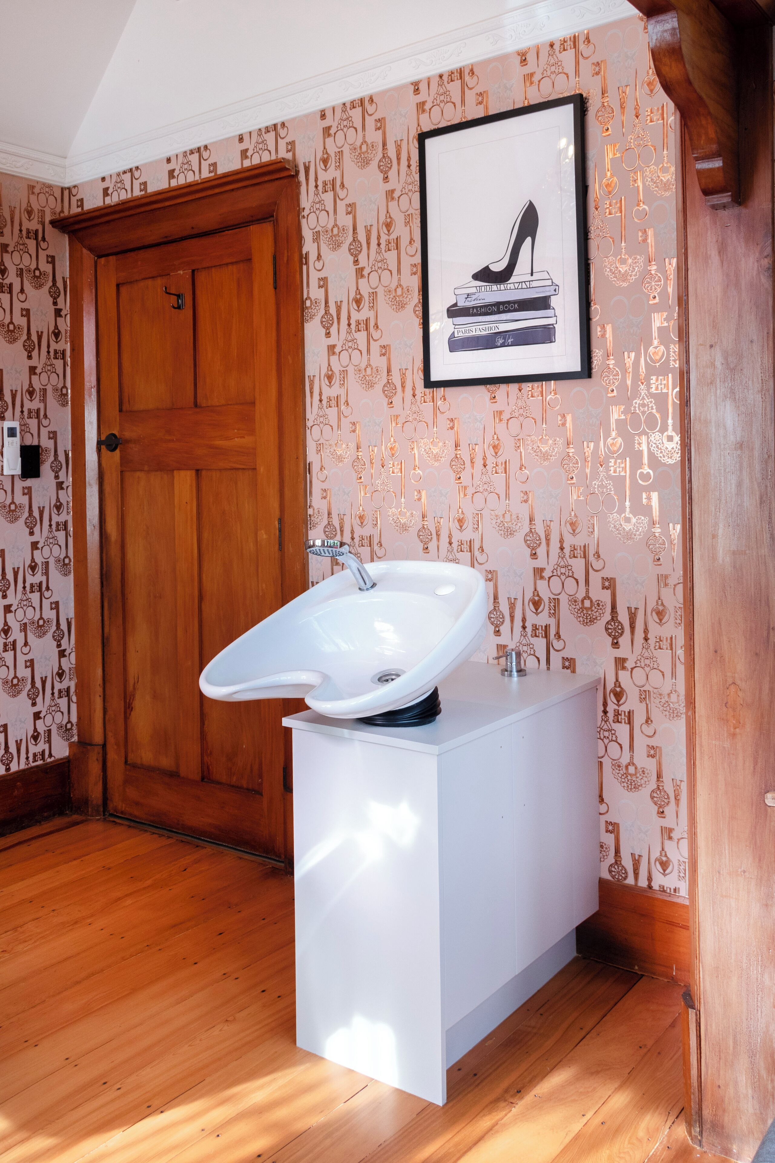 Ein Badezimmer mit Holztür, einem weißen Waschbecken auf Fuß und einem gerahmten Bild von einem High Heel Schuh, das auf Büchern an der Wand angebracht ist.