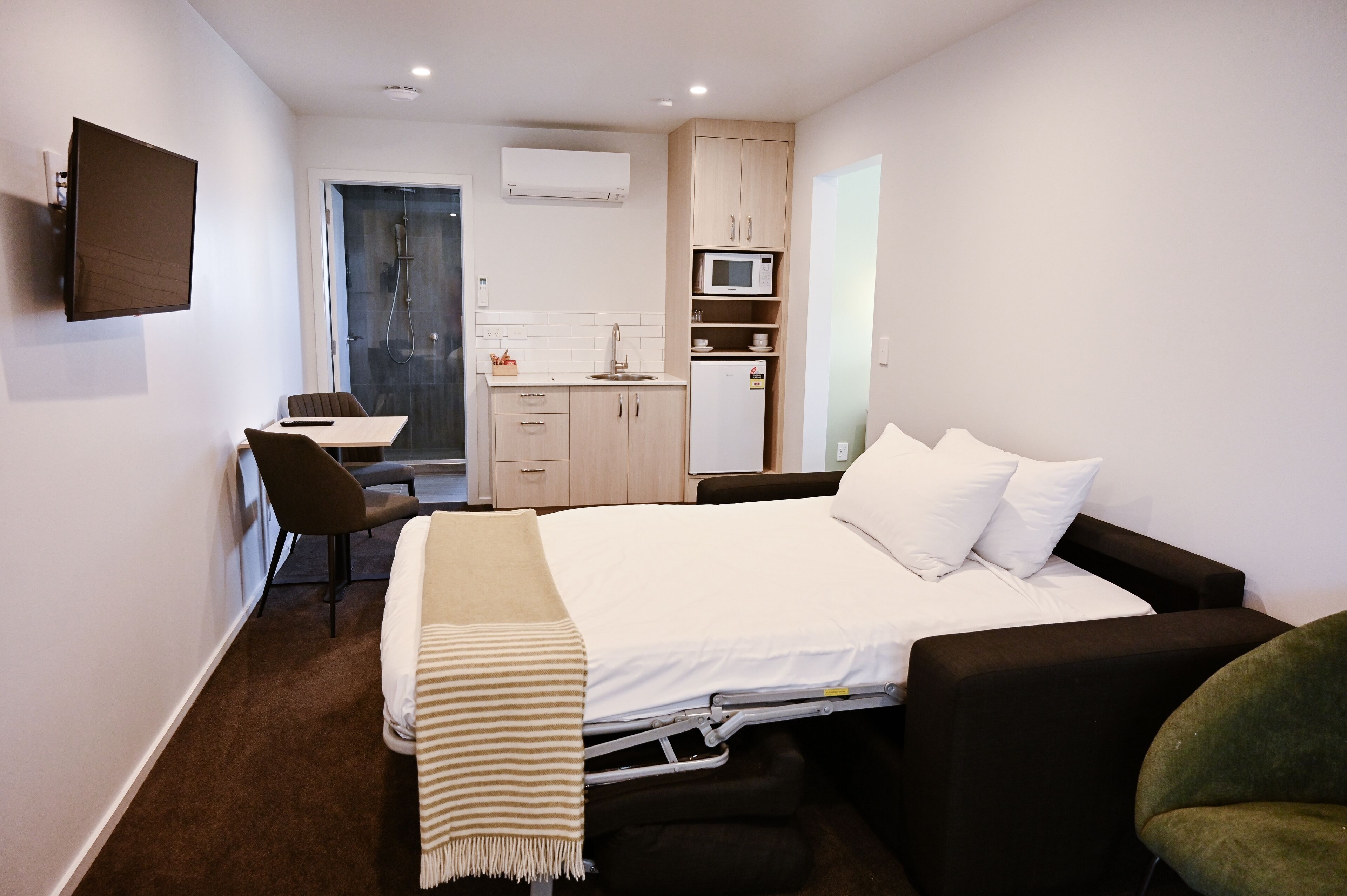 Een compacte hotelkamer met een bed, een bureau met stoel, een kitchenette en een douche.