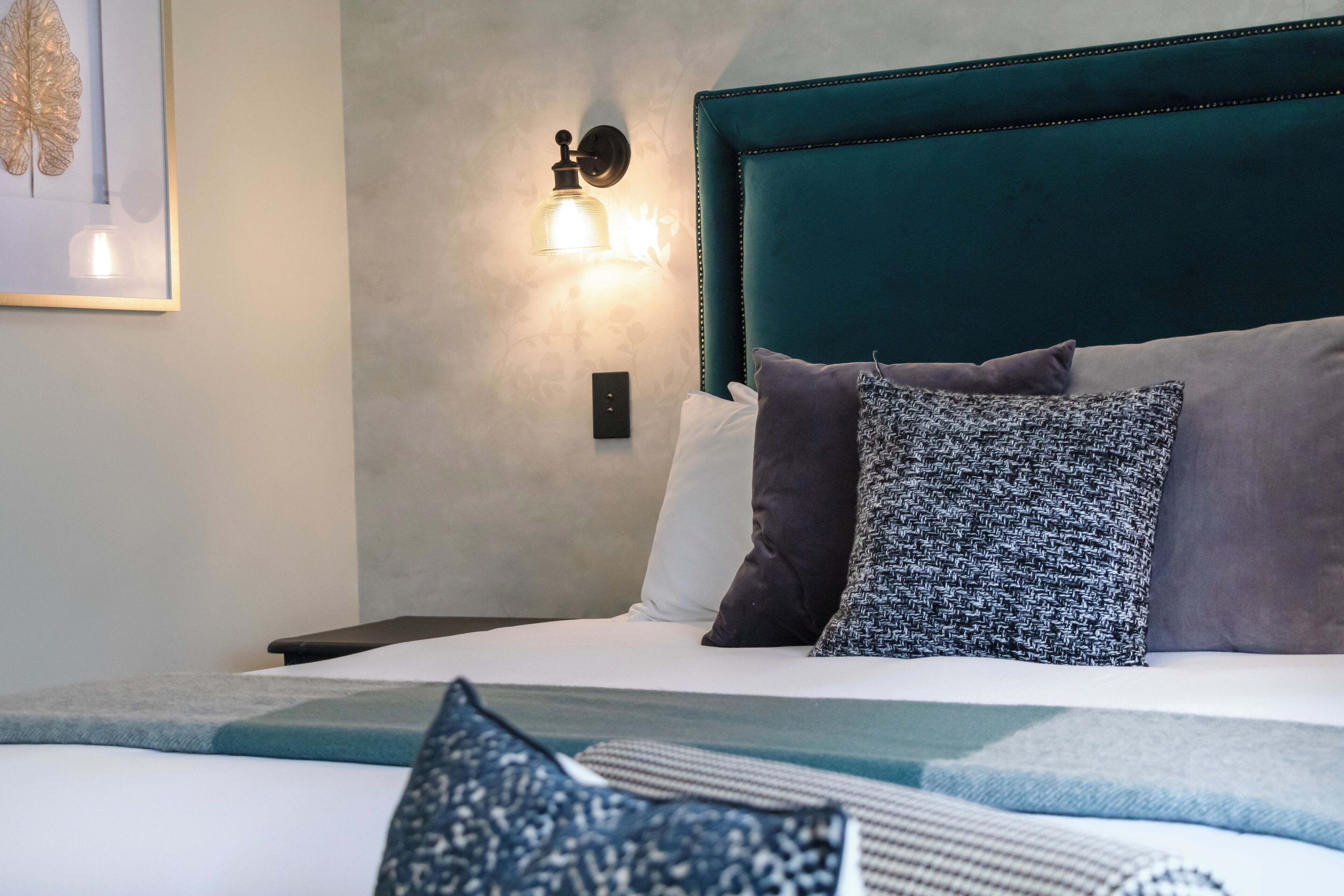 Un lit bien fait, avec une tête de lit de couleur turquoise, plusieurs oreillers et un luminaire fixé au mur.