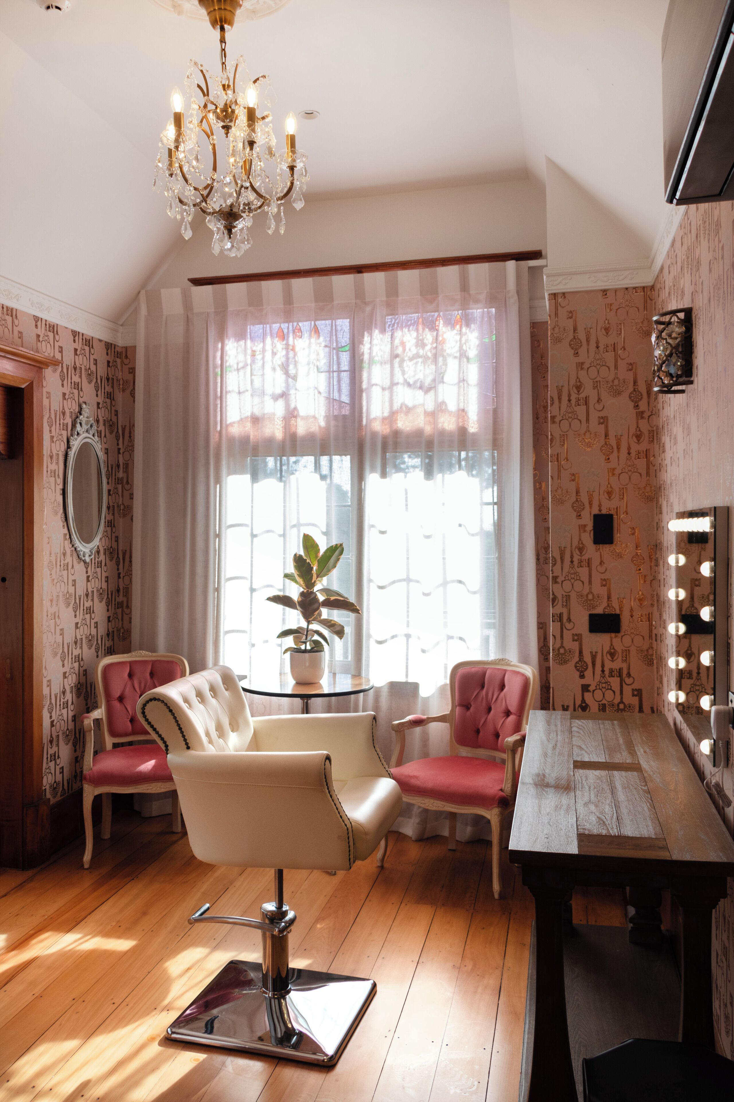 A room with a chandelier, a vanity with lights, two chairs, a table, and a potted plant.
