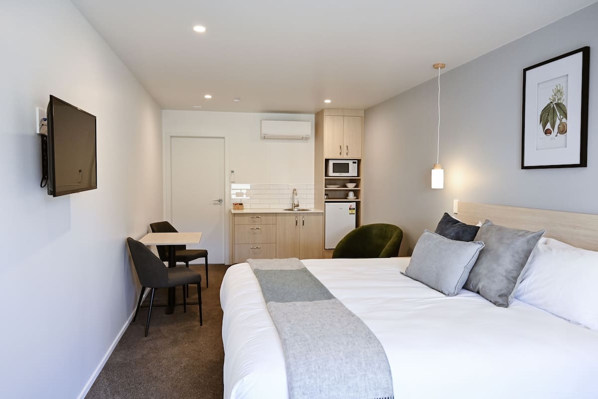 A modern hotel room with a bed, a desk, a chair, a kitchenette, and a television.