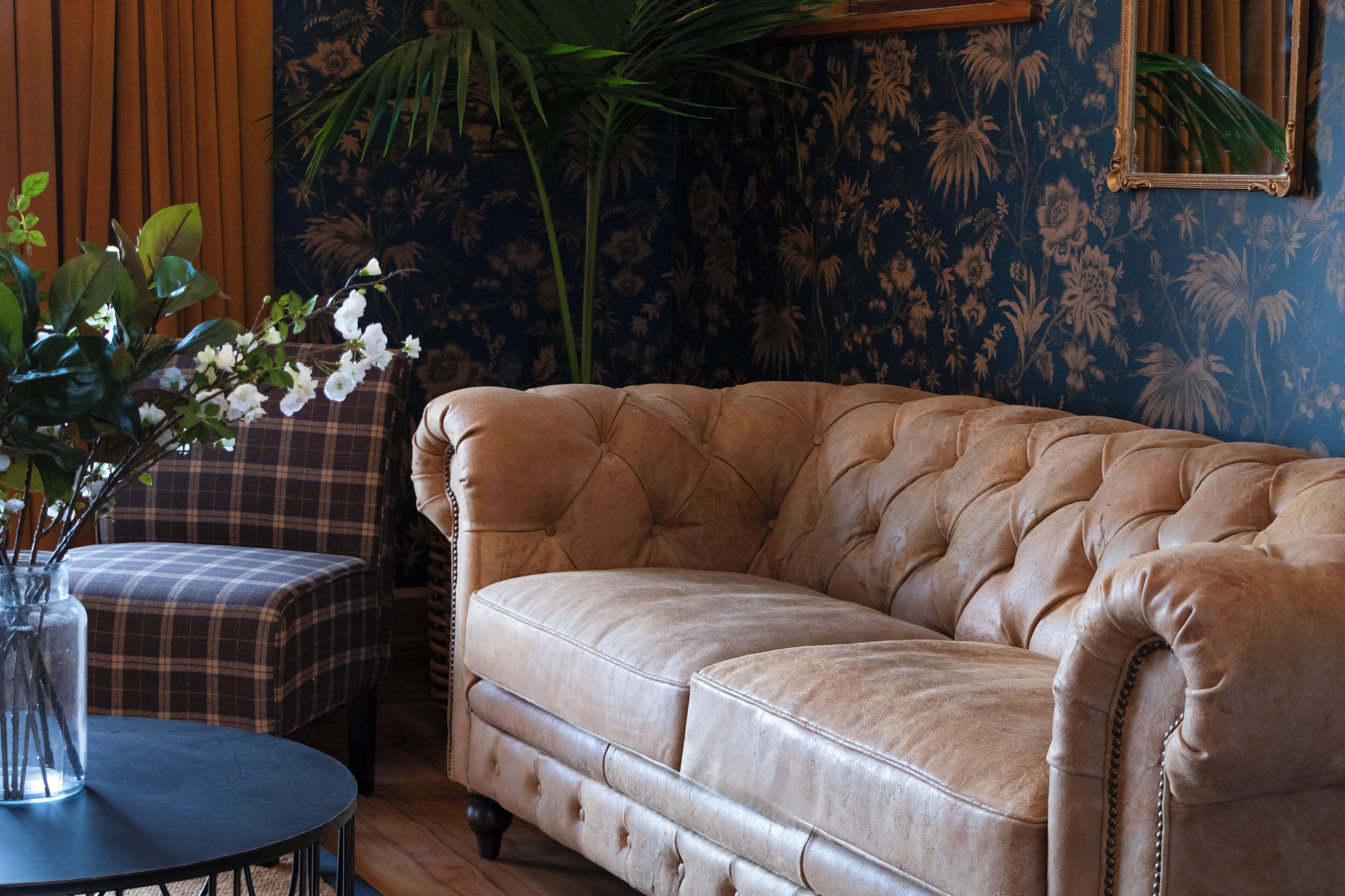Un canapé en cuir capitonné se trouve dans une pièce dont les murs sont recouverts d’un papier peint à motifs floraux. On y aperçoit également un fauteuil à carreaux et une petite table ronde sur laquelle est posé un vase rempli de fleurs.