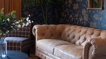 Un canapé en cuir capitonné se trouve dans une pièce dont les murs sont recouverts d’un papier peint à motifs floraux. On y aperçoit également un fauteuil à carreaux et une petite table ronde sur laquelle est posé un vase rempli de fleurs.