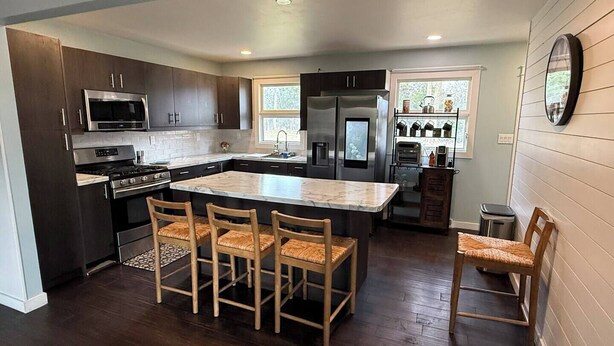 Una cocina moderna con alacenas de madera oscura, electrodomésticos de acero inoxidable, una isla de mármol con cuatro sillas y un ventanal que proporciona luz natural.