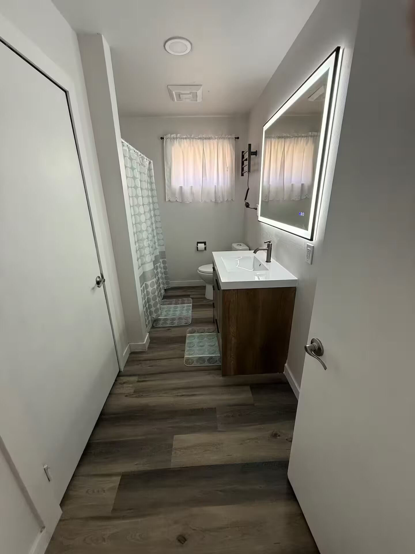 Un couloir étroit mène à une salle de bain équipée d’un meuble-lavabo en bois, de toilettes et d’une douche avec un rideau.