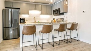 A modern kitchen with a stainless steel refrigerator, a microwave, and a coffee machine. There are four bar stools and a white countertop.