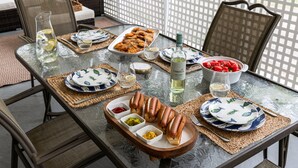 Een eettafel gedekt met hotdogs, sauzen en een fles wijn, omringd door rieten stoelen en een comfortabele bank.