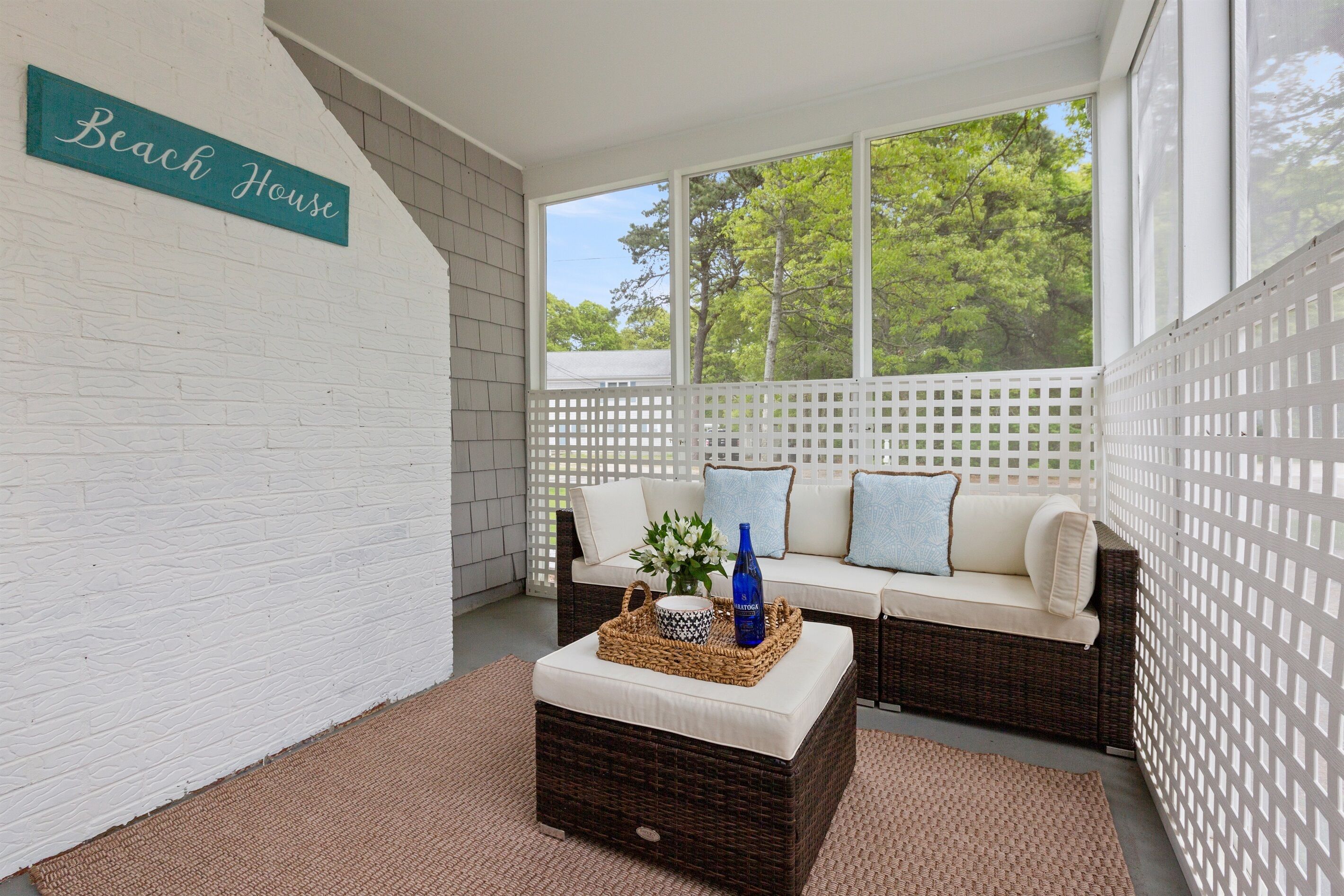 Un área de estar exterior acogedora con mobiliario de mimbre, una pared blanca con un letrero que dice 'Beach House' y vistas a árboles a través de amplias ventanas.