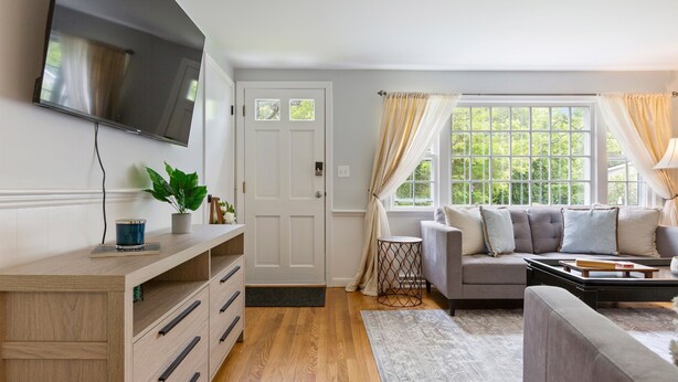 Een woonkamer met een flatscreen televisie aan de muur, een bank met kussens, een salontafel, een houten kast met lades en een potplant.