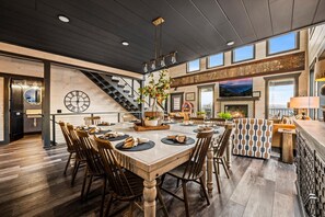 Ein geräumiger Essbereich mit einem großen Holztisch, mehreren Stühlen und einer zentralen Vase mit Blumen. Im Hintergrund ist eine Treppe zu einer oberen Ebene zu sehen.