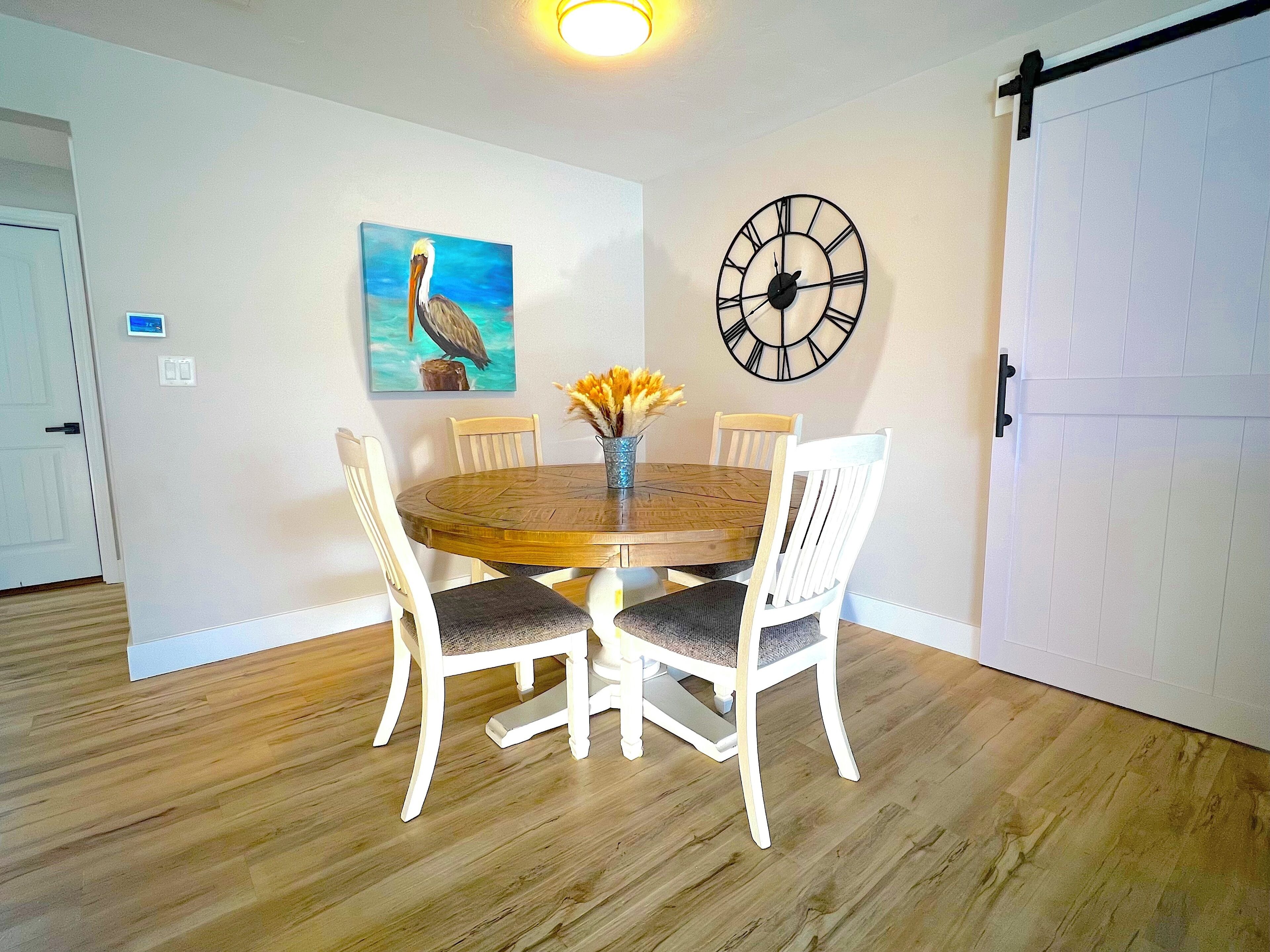 Un coin repas avec une table en bois, des chaises blanches, une horloge et un tableau représentant un oiseau.