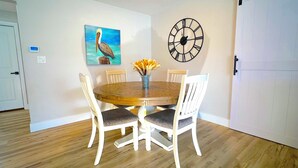 Uma área de refeições com uma mesa de madeira, cadeiras brancas, um relógio e uma pintura de um pássaro.