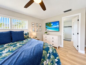 A bedroom with a bed, a nightstand, a dresser, a TV, and a door leading to a bathroom.