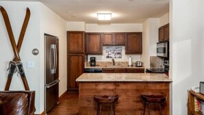 A kitchen with wooden cabinets, a stainless steel refrigerator, a microwave, and a large island with stools. - Modern 2 Bedroom Condo at the Base of Vail Mountain Next to Gondola One (Vail)
